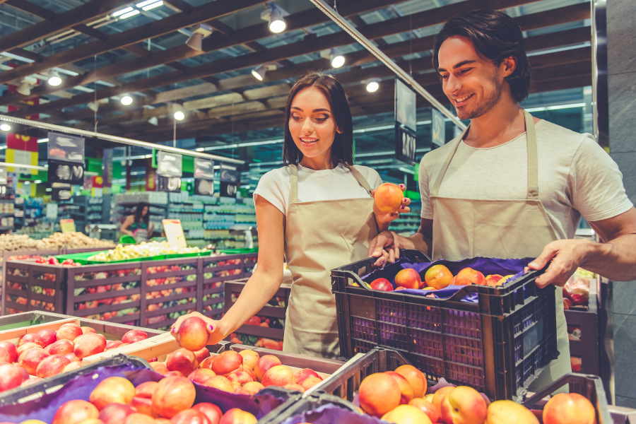 Zwei Mitarbeitende in Arbeitskleidung sortieren gemeinsam frisches Obst in einem gut beleuchteten Supermarkt. Sie wirken freundlich und engagiert.</p>
<p>