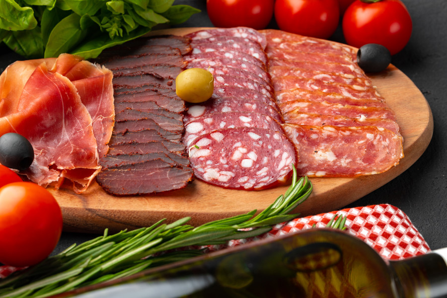 Hochwertige Wurstplatte mit verschiedenen luftgetrockneten Spezialitäten: Parmaschinken, Bresaola, Salami und Coppa – serviert auf einem Holzbrett mit Oliven, frischen Kräutern, Tomaten und grünem Salat im Hintergrund.